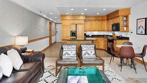 A modern living room with a brown leather sofa, two armchairs, a glass coffee table, and a kitchen area with wooden cabinets and appliances.