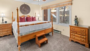 Une chambre à coucher avec un lit à baldaquin, des tables de chevet en bois, une commode, un ventilateur de plafond et une grande fenêtre donnant sur la forêt.