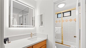 Une salle de bain avec un lavabo blanc, un miroir, des meubles en bois et une baignoire dotée d’un cadre doré.