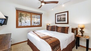 A bedroom with a large bed, a ceiling fan, a window with a view of trees, a framed picture on the wall, and a bedside table with a lamp.