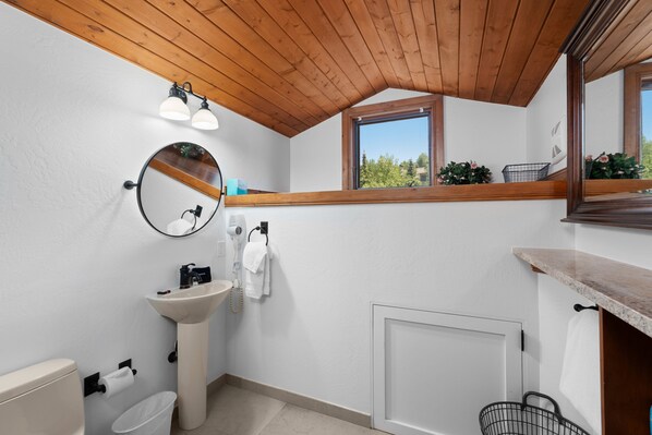 Un baño con techo de madera, un espejo redondo, un lavabo, un inodoro y una ventana con vistas a la vegetación.