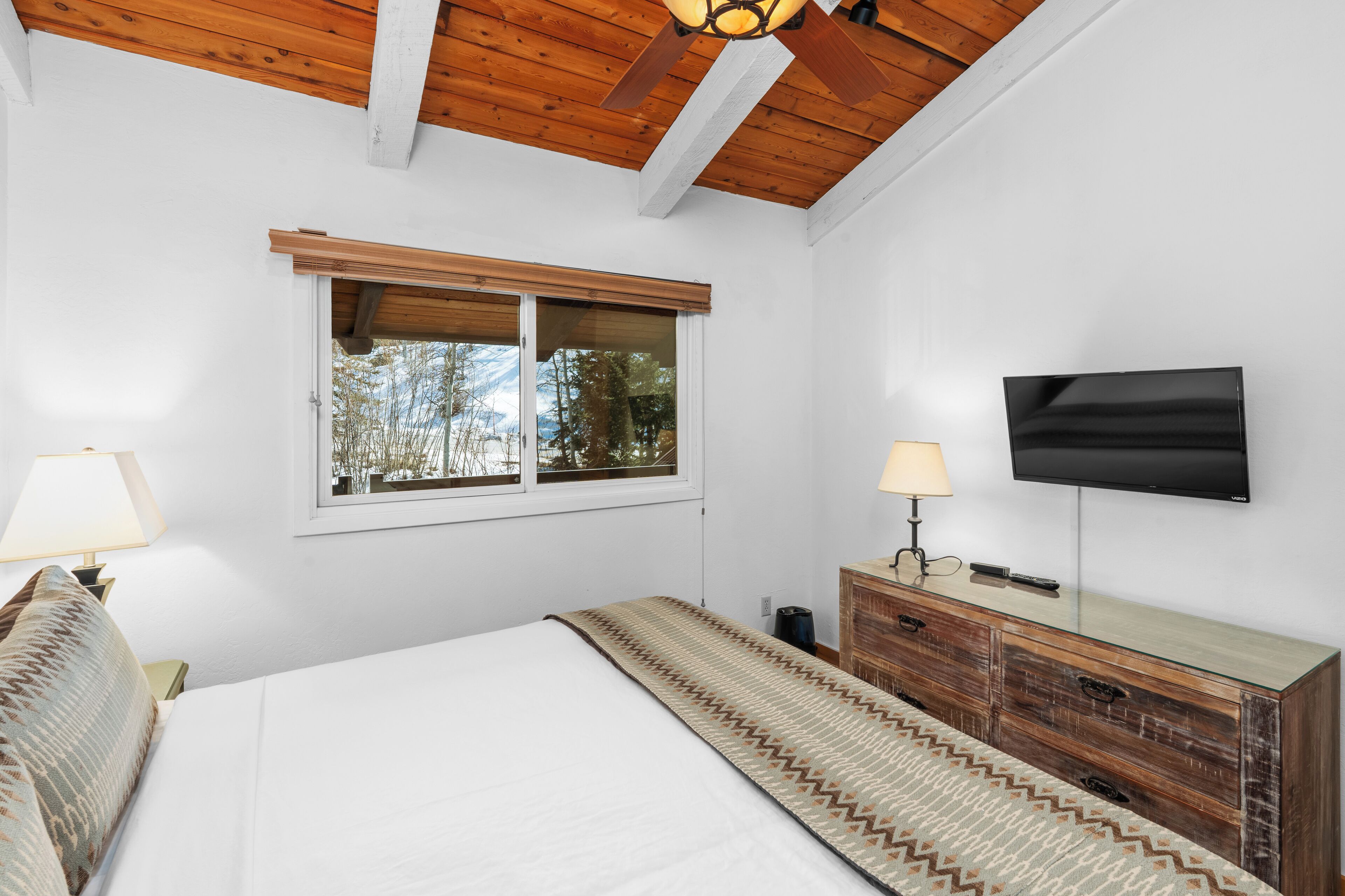 A bedroom with a bed, a wooden ceiling, a window with blinds, a TV, and a dresser.