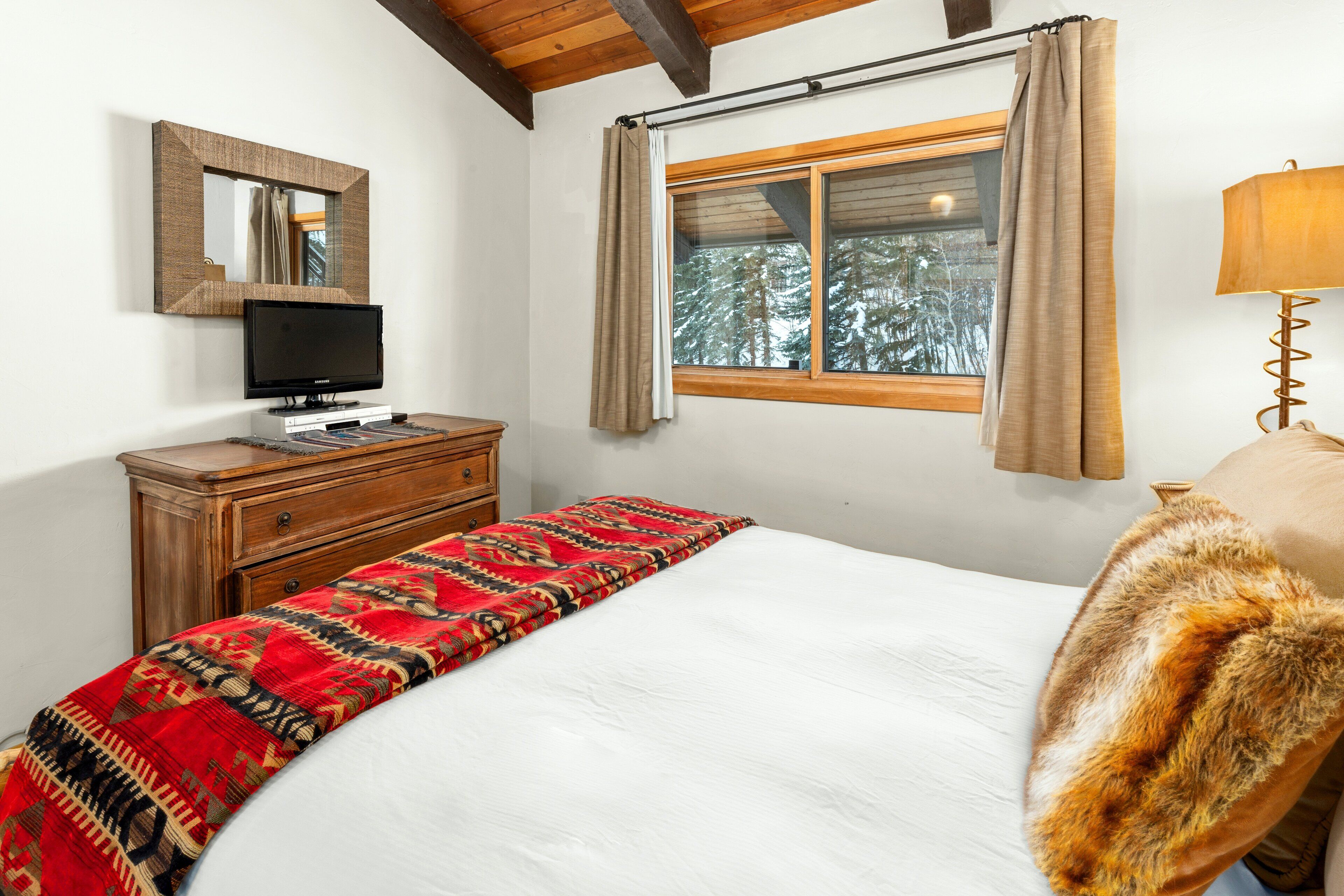 A bedroom with a bed, a wooden dresser, a television, a window with curtains, and a lamp.