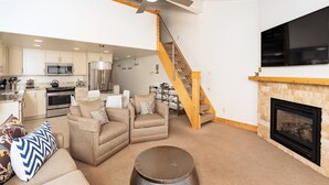 A modern living room with a fireplace, a kitchen in the background, and a wooden staircase. - Crestwood 2307 Premier 1 Bedroom Plus Loft (Snowmass Village)
