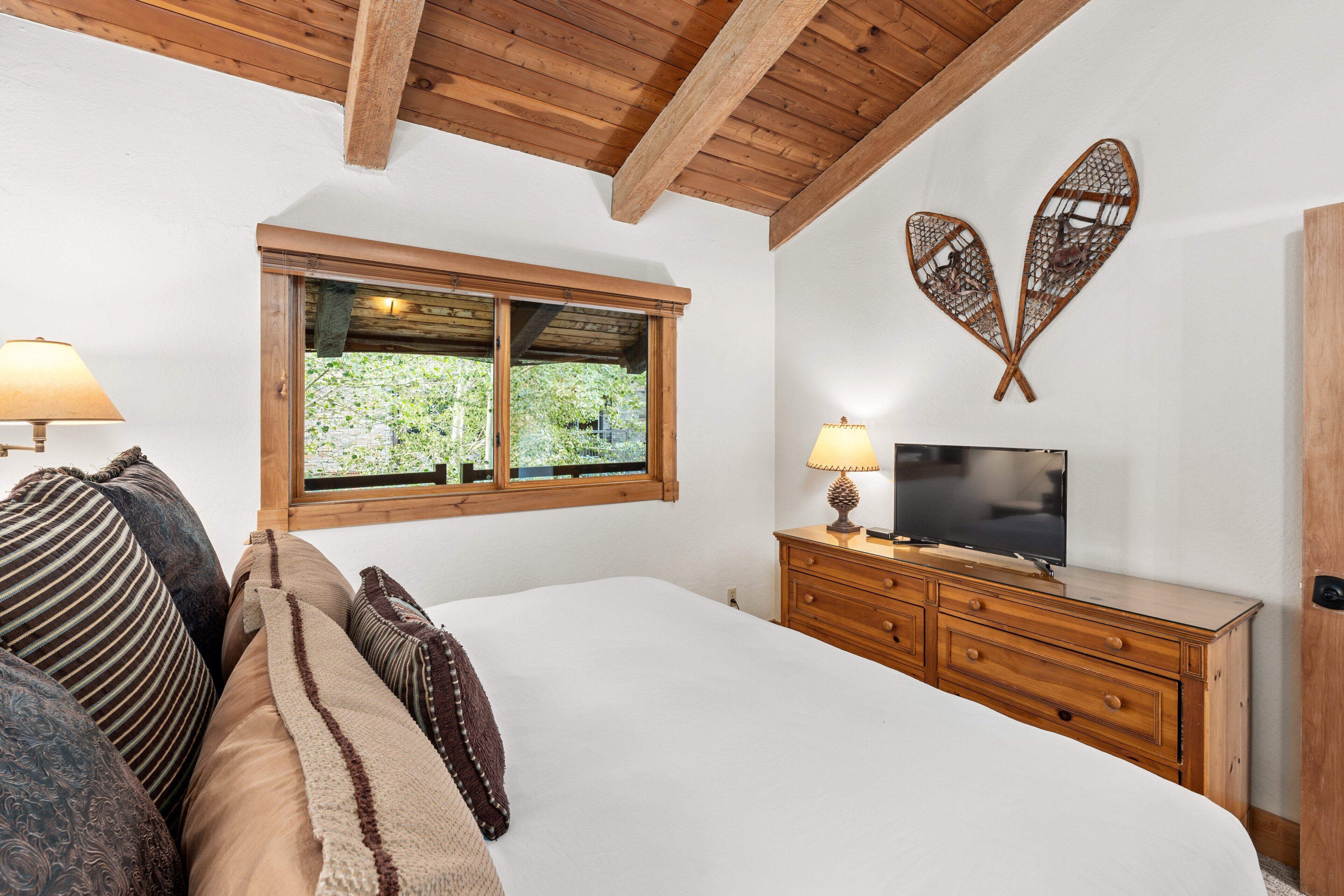 Un dormitorio con techo de madera, una cama con numerosas almohadas, un televisor sobre un aparador de madera y una ventana con vistas a zonas verdes.