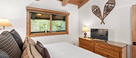 Un dormitorio con techo de madera, una cama con numerosas almohadas, un televisor sobre un aparador de madera y una ventana con vistas a zonas verdes.