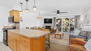 A modern kitchen with wooden cabinets, a central island, and pendant lighting. There is a dining area with a sofa and chairs, and a living room with a fireplace and a large window.