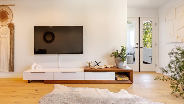 Una sala de estar moderna con un televisor de pantalla plana montado en una pared blanca, una consola de madera con objetos decorativos, una alfombra blanca y una puerta que da a un balcón.