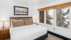 Un dormitorio con una cama grande, dos mesitas de noche con lámparas, un cuadro enmarcado sobre la cama y una ventana con vistas a árboles y un paisaje nevado.