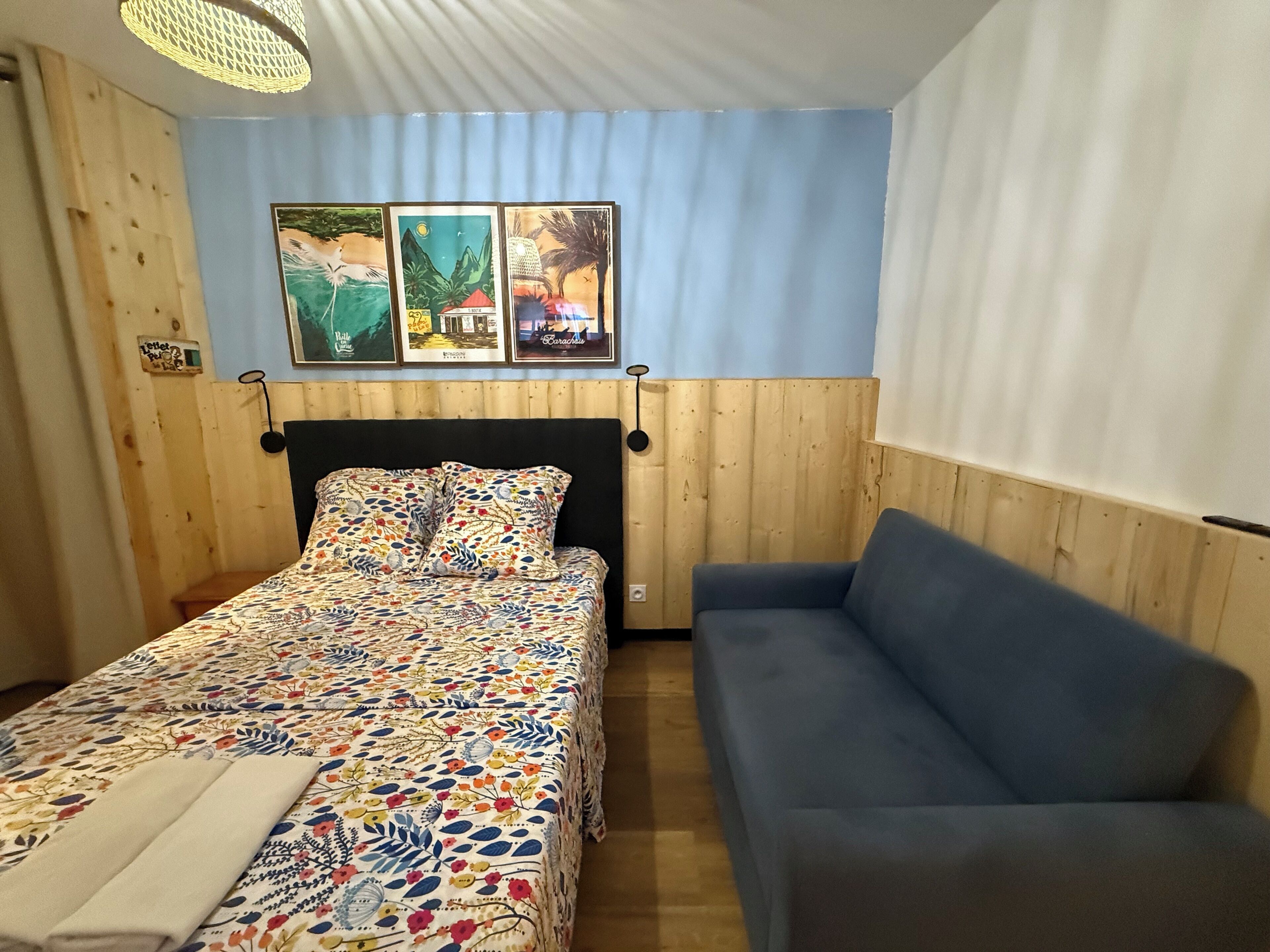 A room with a bed, a blue sofa, wooden walls, and three framed pictures on the wall.