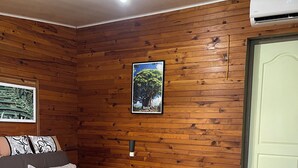 Ein gemütliches Zimmer mit einer Holzwand, einem gerahmten Bild von einem Baum, einer Couch mit Kissen, einem Holztisch und einem Stuhl.