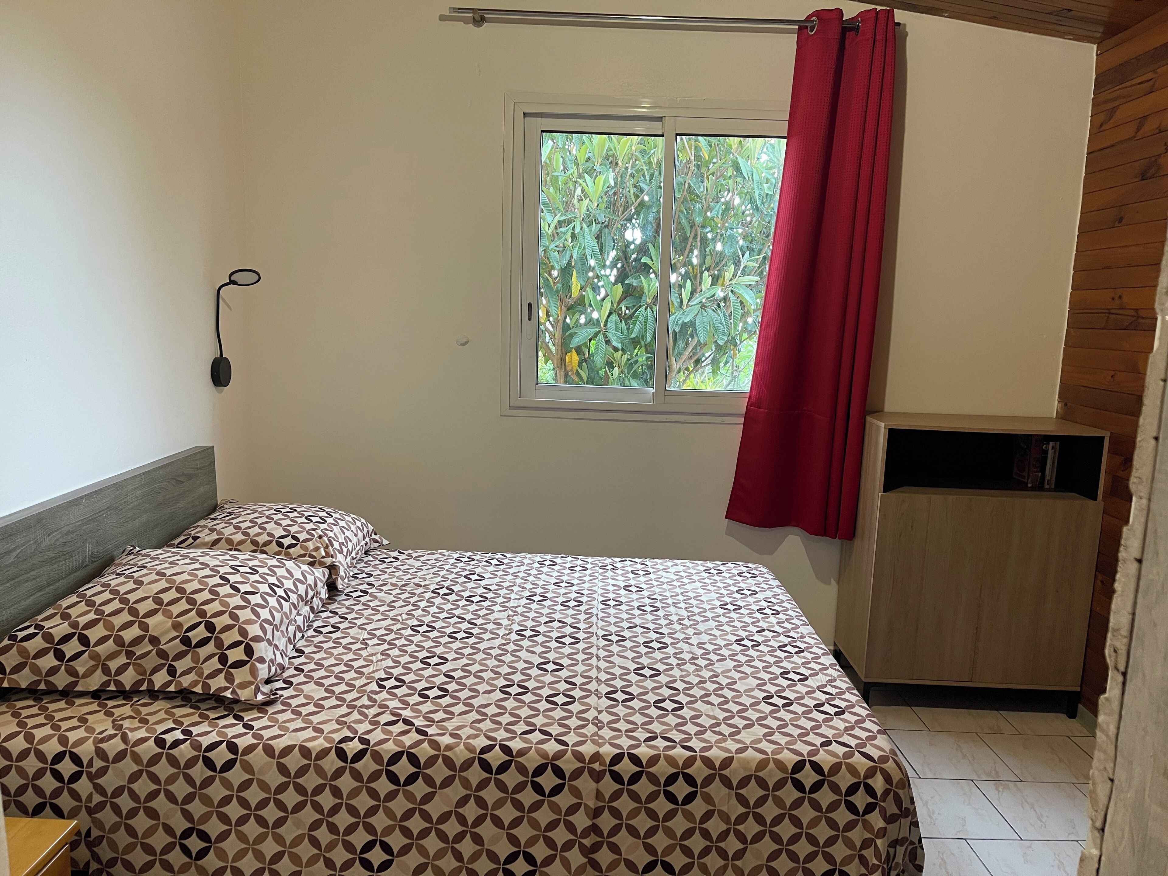 A room with a bed, a window with a red curtain, a wooden cabinet, and a wall-mounted power outlet.