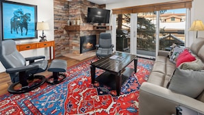 A living room with a stone fireplace, leather chairs, a sofa, a TV, and a large window with a view. - Crestwood 3108 Premier 2 Bedroom (Snowmass Village)