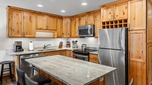 A kitchen with wooden cabinets, stainless steel appliances, a marble countertop, and black bar stools. - Crestwood 3109 Deluxe 2 Bedroom (Snowmass Village)