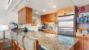 A kitchen with a granite countertop, wooden cabinets, a stainless steel refrigerator, and a bar area with cushioned chairs.