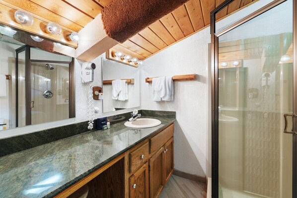 Une salle de bain dotée d’une douche à parois de verre, d’un lavabo avec un plan de travail en marbre, de meubles en bois et d’un grand miroir avec des lumières au-dessus.