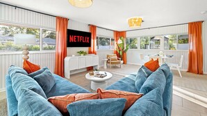 A modern living room with a large TV screen displaying 'NETFLIX', a sofa set, a coffee table, and a dining area with a white table and chairs.