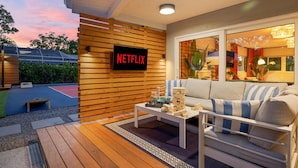 A modern living room with a Netflix sign, a wooden deck, and a view of a tennis court.
