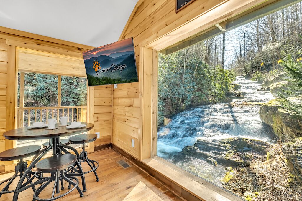 Ein Holzhütten-Interieur mit Tisch und Stühlen, einem großen Fenster mit Blick auf einen Wasserfall und einem Poster an der Wand.