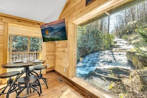 Ein Holzhütten-Interieur mit Tisch und Stühlen, einem großen Fenster mit Blick auf einen Wasserfall und einem Poster an der Wand.