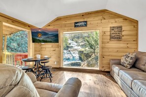 A cozy cabin interior with a wooden ceiling, a large window overlooking a waterfall, a dining area with a table and chairs, and a sofa. - Smoky Mtn Cabin w/Creek Views and Waterfall (Cosby)