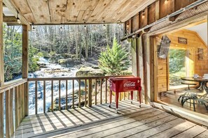 A wooden deck with a view of a river and forest, a Coca-Cola cooler, and a dining area with outdoor seating.