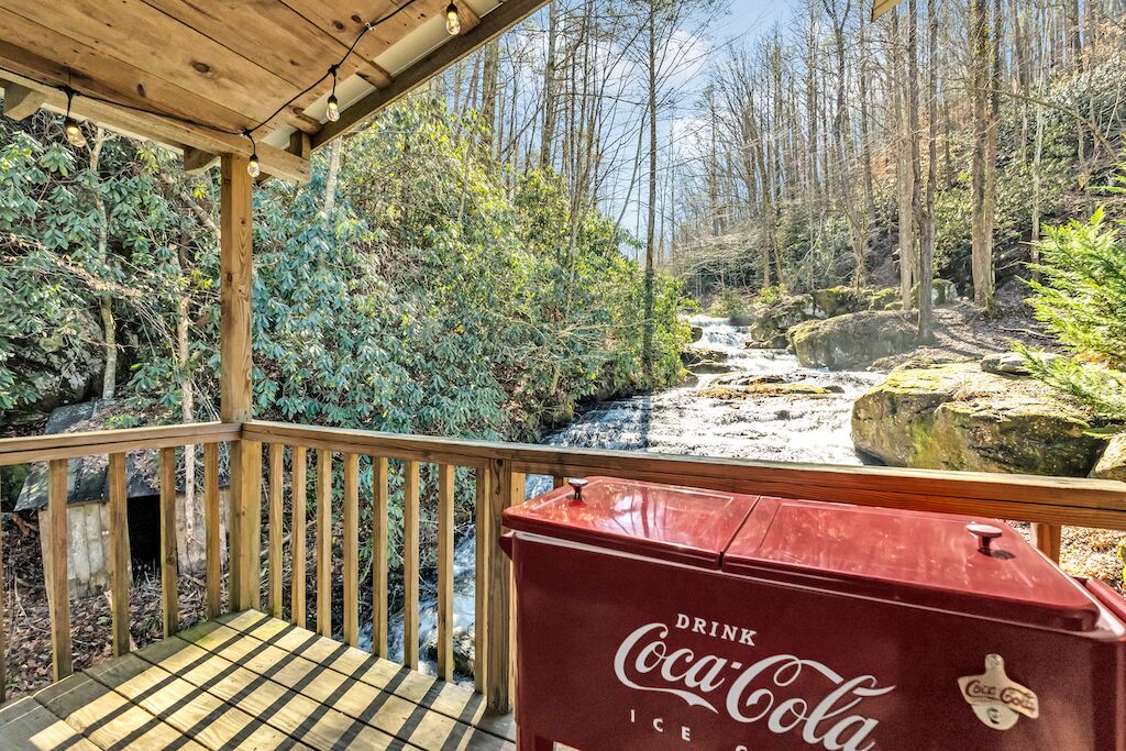 Una terraza de madera con un enfriador de hielo con la marca Coca-Cola, con vistas a una zona boscosa con un arroyo.
