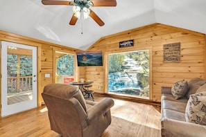 Interior acogedor de una cabaña con techo de madera, ventilador de techo, sofá, silla, mesa y un amplio ventanal con vista a una cascada.