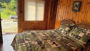 Ein Schlafzimmer einer Holzhütte mit einer gesteppten Bettdecke, einem Fenster mit Jalousien und einer Tür, die nach draußen führt.