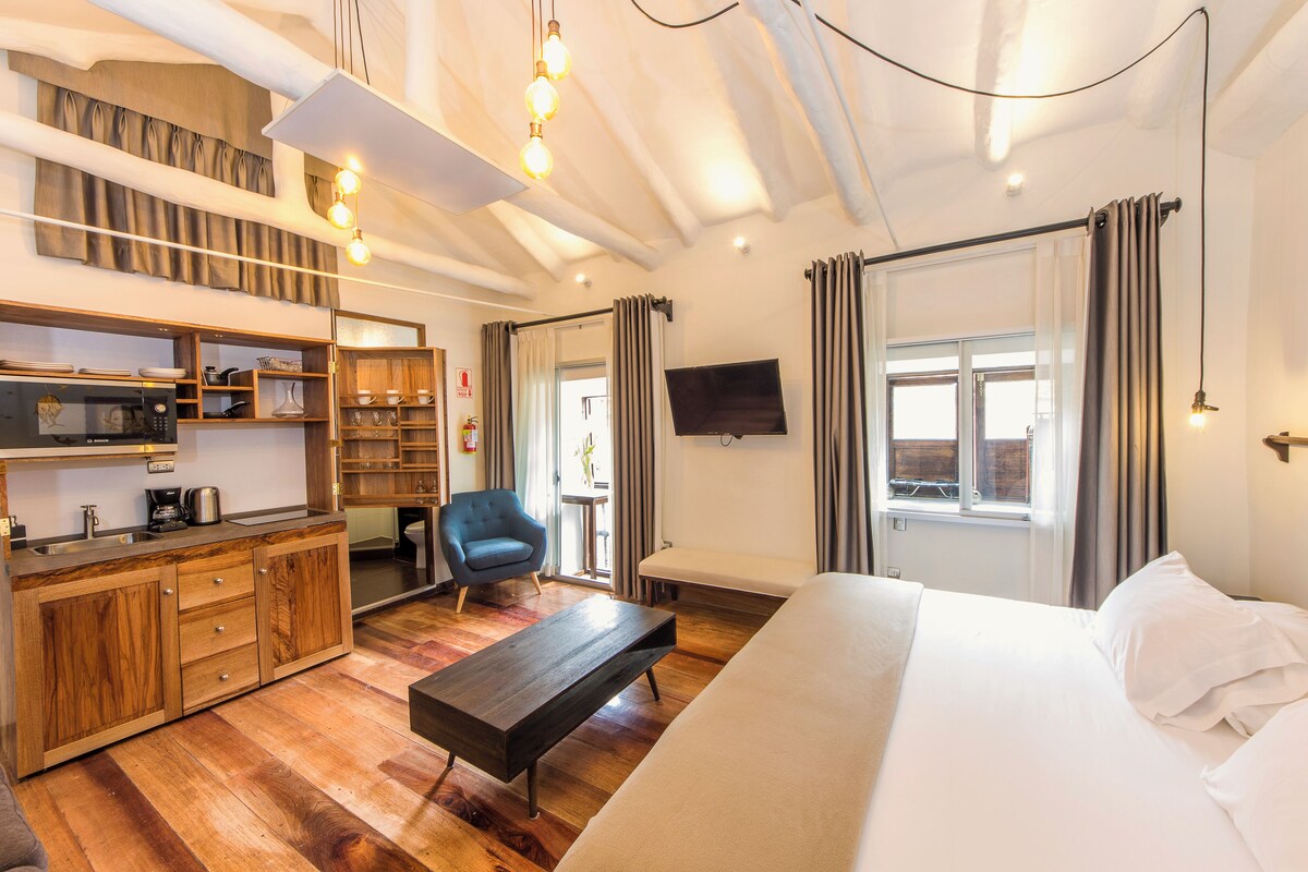 Une chambre d’hôtel moderne avec une kitchenette en bois, un grand lit, un banc et un téléviseur fixé au mur.