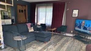 A living room with a grey sofa, a small table, a TV displaying a news channel, and a window with curtains. - Charming Red Tower, Loft  ambiance, Mountain views, AC, WiFi, Berkshires NY (Petersburgh)