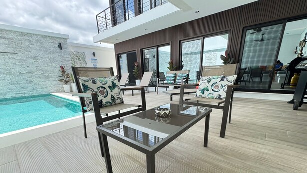 Ett modernt utomhusområde med sittplatser, ett glasbord och stolar, en pool och utsikt över havet.