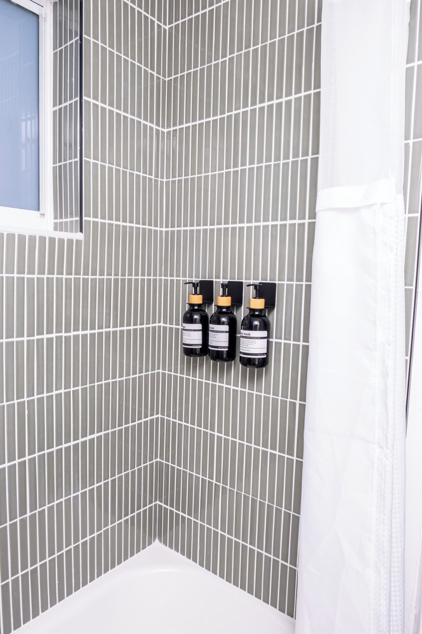 Un baño con pared de azulejos, una bañera blanca y tres botellas negras fijadas a la pared.