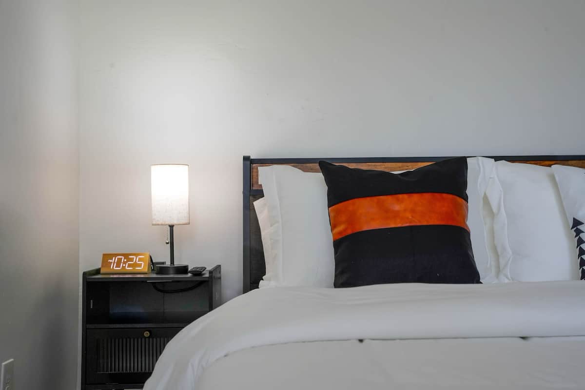 Ein ordentlich bezogenes Bett mit weißer Bettwäsche, einem schwarzen und orangefarbenen Kissen sowie einem Nachttisch mit Lampe und digitaler Uhr, die 10:25 anzeigt.