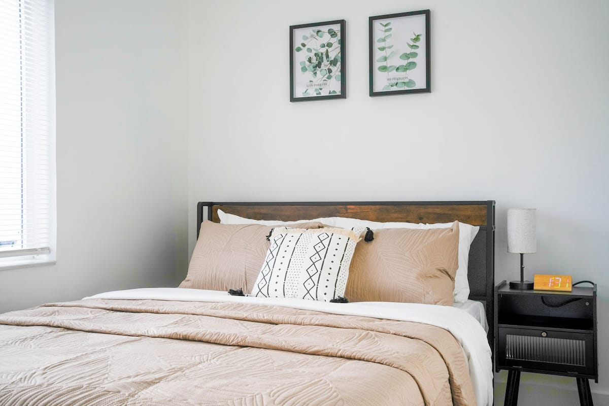 Ein ordentlich gemachtes Bett mit einer beigen Tagesdecke und Kissen, ein Nachttisch mit einer Lampe und einer Uhr, und zwei gerahmte botanische Drucke an der Wand.