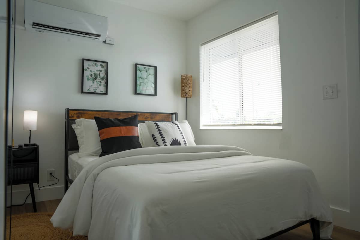 Ein Schlafzimmer mit einem Bett, einem Nachttisch, einer Klimaanlage und einem Fenster mit Jalousien.