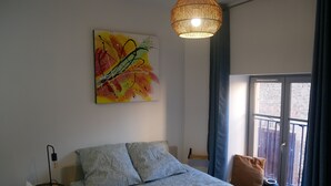 A bedroom with a bed, a bedside table with a lamp, a window with curtains, and a wall-mounted abstract painting. - Cosy studio in the heart of the village of Chazay (Chazay-d'Azergues)