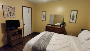 Ein Hotelzimmer mit einem Bett, einem Fernseher auf einem Holzständer, einem Schminktisch und zwei gerahmten Blätter-Kunstwerken an der Wand.