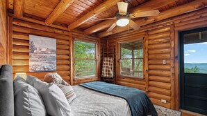 A log cabin bedroom with a bed, a ceiling fan, and a large window offering a view of the outdoors.
