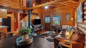 A log cabin interior with a wooden dining table, a leather sofa, a TV, and a fireplace.