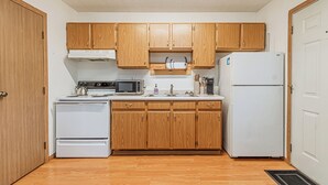 A compact kitchen with wooden cabinets, a white stove, a microwave, and a refrigerator. - 10 min to Sanford I Smart TV I Queen I Free Prking (Fargo)