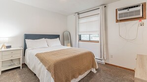 A bedroom with a bed, a nightstand, a bedside lamp, a window with blinds, and an air conditioning unit mounted on the wall. - 10 min to Sanford I Smart TV I Queen I Free Prking (Fargo)