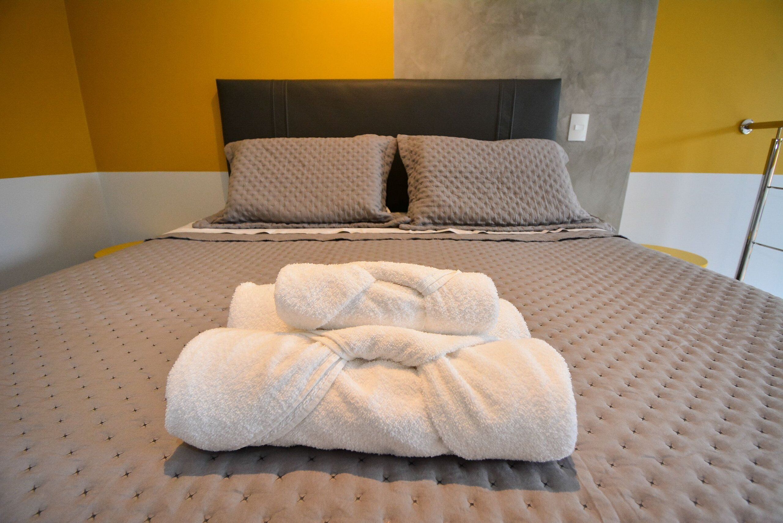 A neatly made bed with a grey duvet and pillows, a folded towel, and a headboard against a yellow and grey wall.