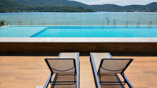 A pool with a view of a lake and mountains, two lounge chairs, and a wooden deck. - Sunset Lagoa (Florianopolis)