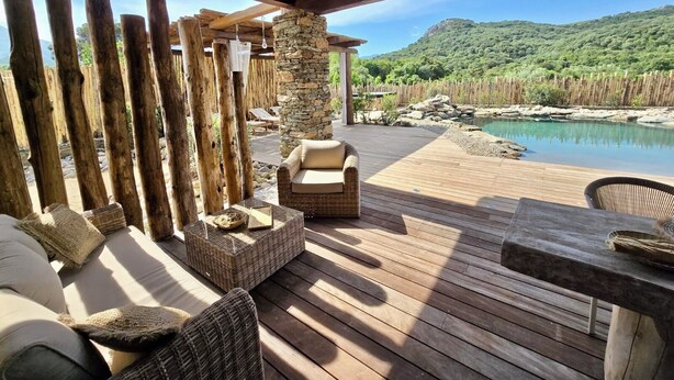A wooden deck with wicker furniture, a stone pillar, and a view of a mountain and pool.