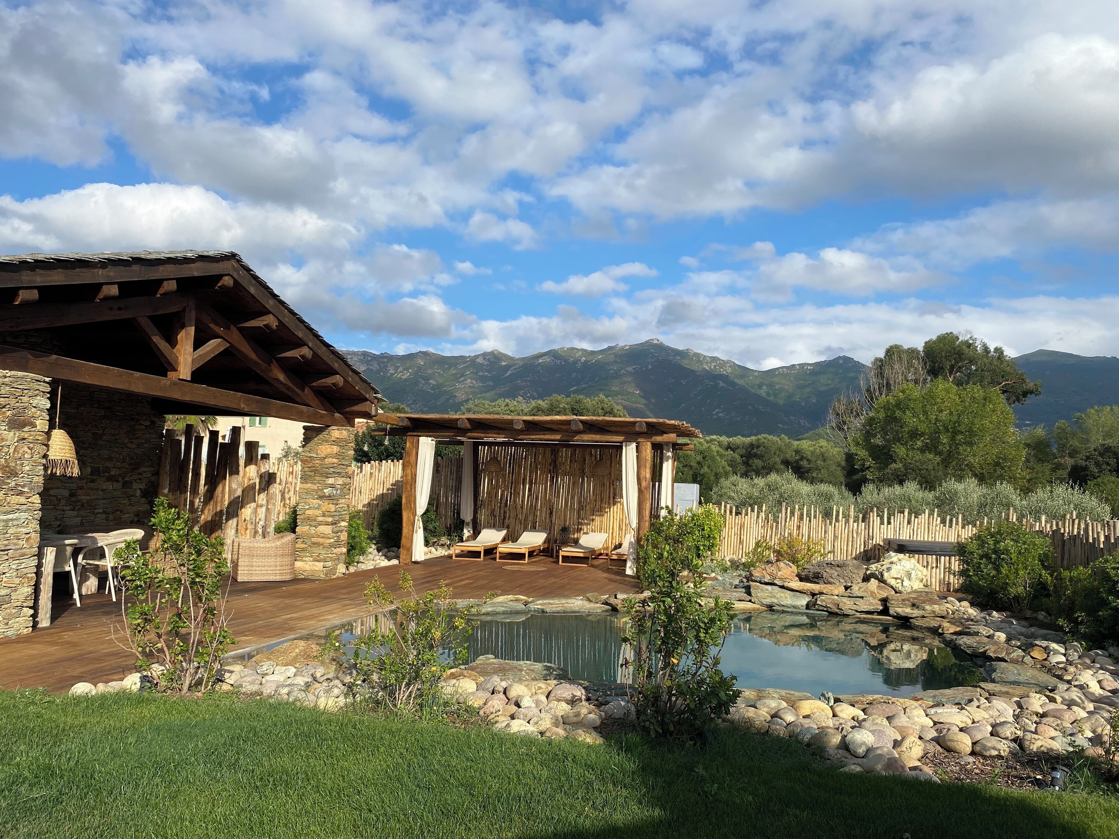 A rustic outdoor seating area with a stone wall, wooden pergola, and a small pond.
