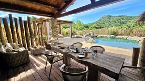 An outdoor dining area with a wooden deck, wicker furniture, and a view of a natural landscape with a pool.
