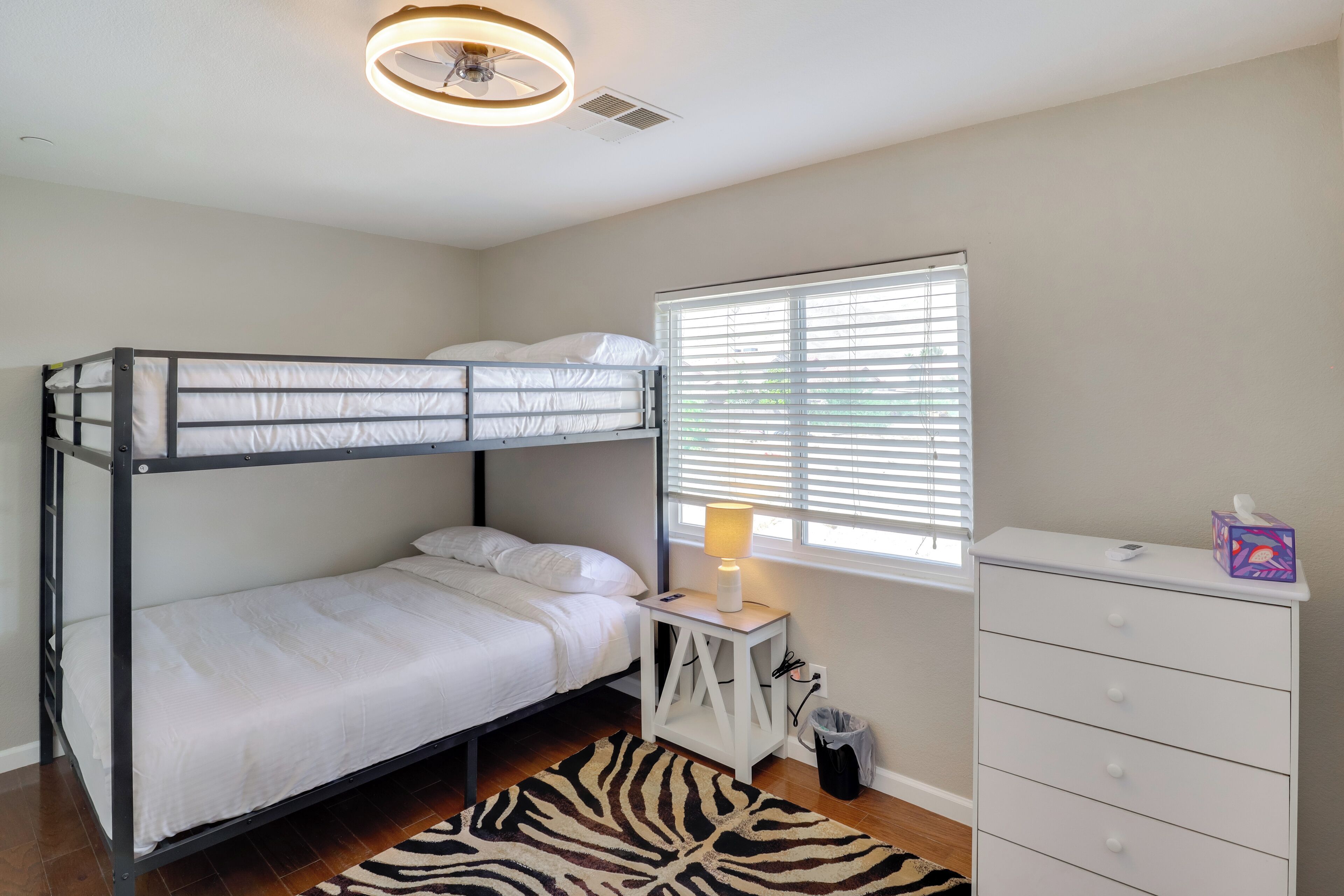A bunk bed room with a white dresser, a zebra-patterned rug, and a window with blinds.