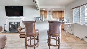 A spacious living area with a fireplace, a flat-screen TV mounted above it, and a kitchen area with wooden cabinets and a window with blinds. - 8 Mi to Dtwn: Home w/ Fireplace & Games in LA! (Los Angeles)
