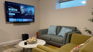 Olohuoneessa on harmaa sohva, kuviollisia koristetyynyjä, koriste-esineillä koristeltu sohvapöytä ja litteänäyttöinen televisio, jolta näkyy sarja nimeltä ”The Last of Us”.